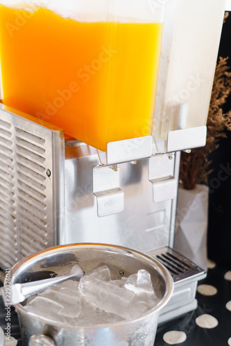 Fresh orange juice and drinking water in beverage dispenser with ice cubes bucket and glass for party on table.