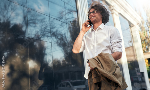 Image of handsome businessman leaning on a building's wall while standing outdoors and using smartphone. Man in suit wearing round spectacles talking with clients at his mobile phone.