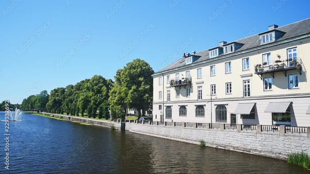 Park and building along the Lidan river in Lidkoping, Sweden, on a sunny summer day. Fountain in the water. Park and building along the Lidan river in Lidkoping, Sweden, on a sunny summer day. Fountain in the water.