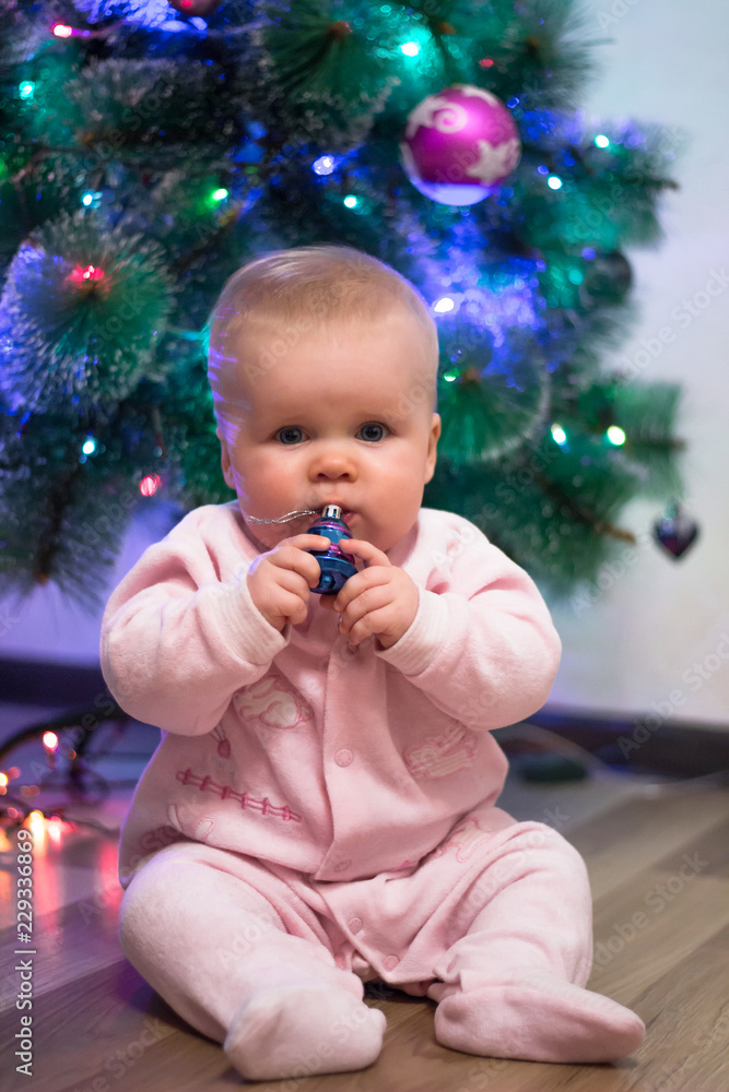 A little girl in a pink jumpsuit is sitting on the background of a Christmas tree, holding a Christmas toy. New Year's holidays. Christmas Holidays. A little girl in a pink jumpsuit is sitting on the background of a Christmas tree, holding a Christmas toy. New Year's holidays. Christmas Holidays.