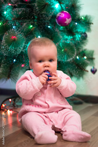 A little girl in a pink jumpsuit is sitting on the background of a Christmas tree, holding a Christmas toy. New Year's holidays. Christmas Holidays.