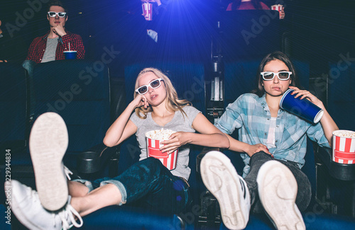 Cool and relaxed girls are sitting in a front row and watching movie. They have put their legs up. Also they are holding their heads with hands. A boy sitting behind them is watching movie as well.