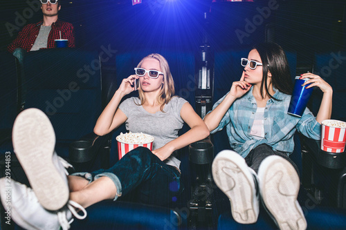 Beautiful and attractive girls are sitting in chairs. Blonde girl is talking on thhe phone. Her friend is showing the silence symbol. She wants her to be quiet and stop talking during watching movie.