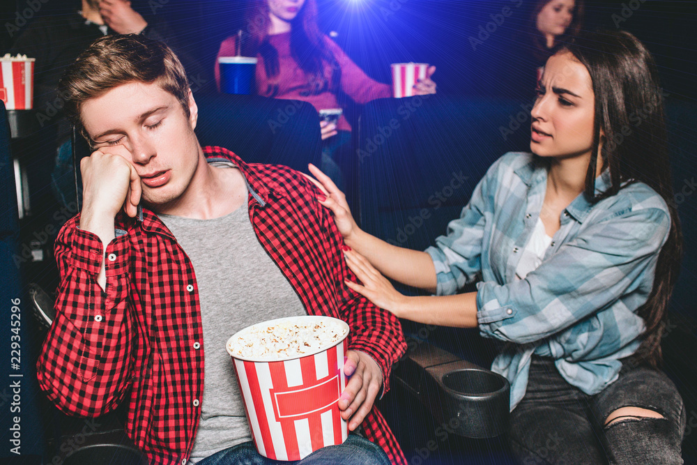 Naklejka premium Guy is sitting in chair and sleeping. He is bored and tired. His girlfriend is trying to wake him up. She is holding it to the hand. Girl is irritated and dissapointed.
