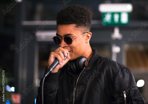 Black rapper performing with microphone. Slight smile. Headphones around neck.