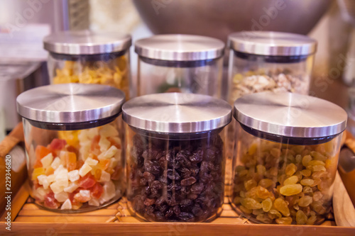 sweet colorful glass mugs, jars or bottles on display in wooden tray in kitchen or pantry for toppings (jelly, currants, sultanas, raisins, grapes, cereal) or ingredients of dessert, snack, ice cream.