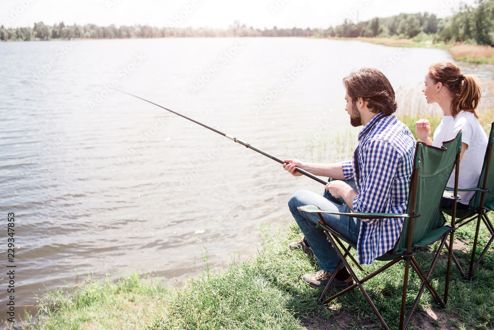 A picture of adult man and woman sitting at the river shore in this nice and beautiful sunny day. Guy is trying to catch some fish. Both of them are looking to the water. A picture of adult man and woman sitting at the river shore in this nice and beautiful sunny day. Guy is trying to catch some fish. Both of them are looking to the water.