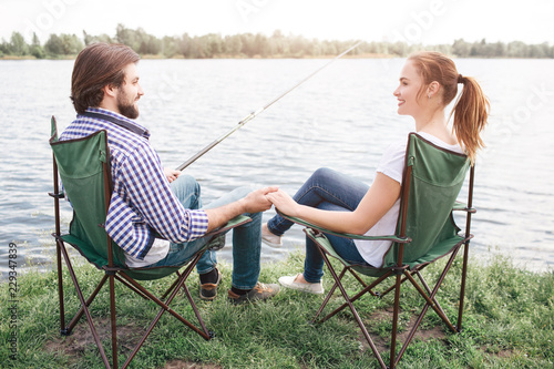 Lovely couple is sitting in soft chairs near river shore and looking at each other. Guy is holding girl's hand. and holding a fish-rod with other one. They look happy.
