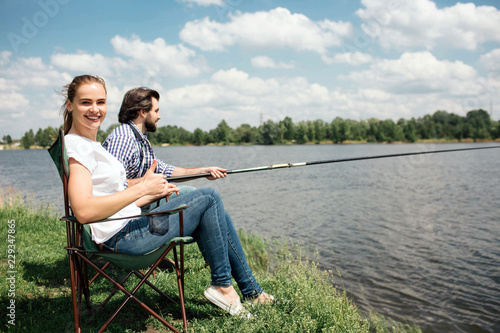 Happy woman is sitting in soft chair and looking on camera. She is smiling. Her husband is sitting besides her and trying to catch some fish using fish-rod.