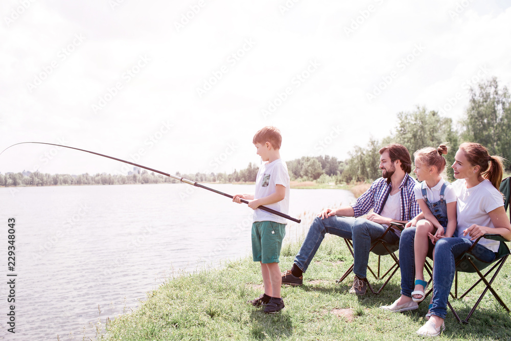 Boy is standing at the edge of river shore and holding fish-rod in hands. It is really long. His parents and sister are sitting behind them and watching it. Boy is standing at the edge of river shore and holding fish-rod in hands. It is really long. His parents and sister are sitting behind them and watching it.