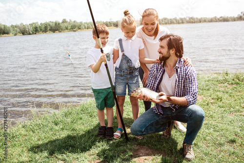 A picture of whole family standing on the grass and looking at fish that dad has caught. Kids and woman look amazed. They are happy. Man is lookiing at them and smiling.