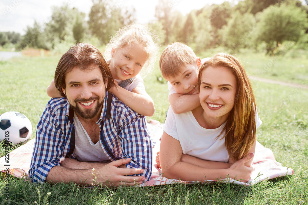 Portrait of family lying on blanket in the middle of meadow. They are looking on camera and smiling. There is a boy lying on woman's back while girl is doing the same thing on man's back. Portrait of family lying on blanket in the middle of meadow. They are looking on camera and smiling. There is a boy lying on woman's back while girl is doing the same thing on man's back.