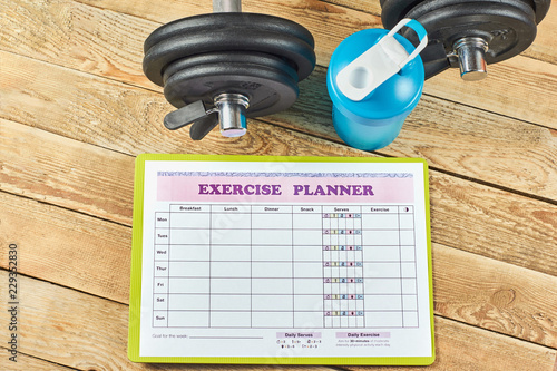 Healthy lifestyle concept. Mock up on workout and fitness dieting diary. Exercise panner sheet, blue shaker and dumbbells on a rustic wooden table