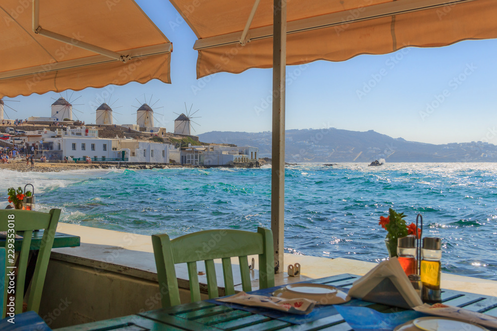  Mykonos, panoramic view of white famous windmills  Chairs with tables in typical Greek restaurant  of Little Venice in Cyclades island, Greece.  
