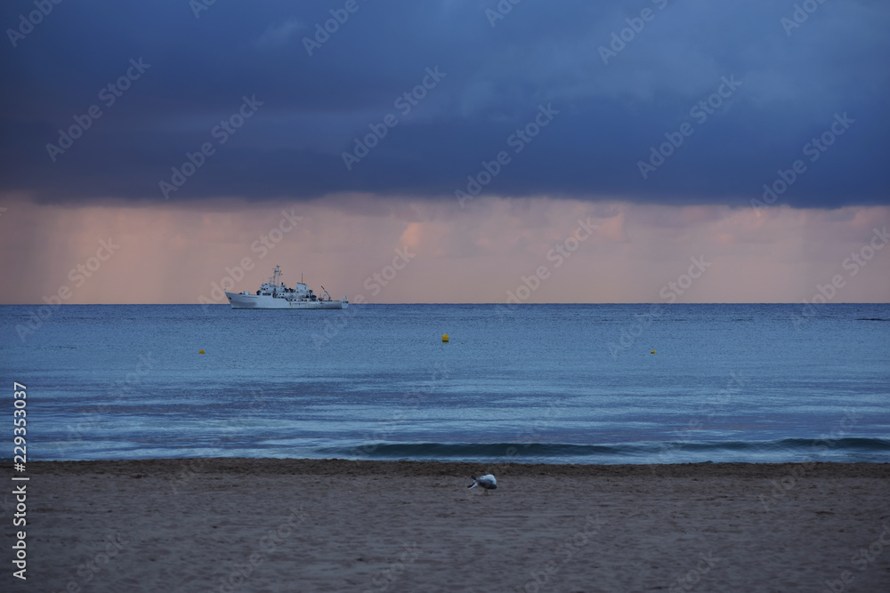 Sandy beach of the Mediterranean coast of Spain with parasols, palm trees and a view of a sailing ship away in cloudy rainy weather