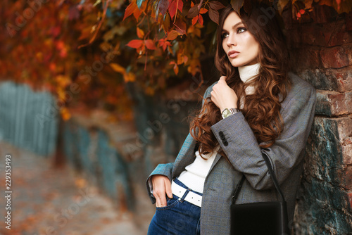 Outdoor fashion portrait of young beautiful fashionable woman wearing white turtleneck, grey checked blazer, wrist watch, holding small leather bag, posing in autumn street. Copy, empty space for text