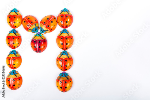 Flat lay with the letter M formed by a group of ceramic ladybugs with red, orange, yellow, blue and black colors on a white studio background with space for text