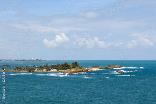 View of a tropical island in the blue sea, surrounded by reefs. Blue sky with clouds. Palm trees and beach. Waves on stone reefs.