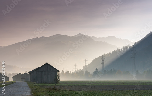 morning mist in a mountain valley with fields and old wooden barns and lattice cross power lines and mountains in silhouette behind