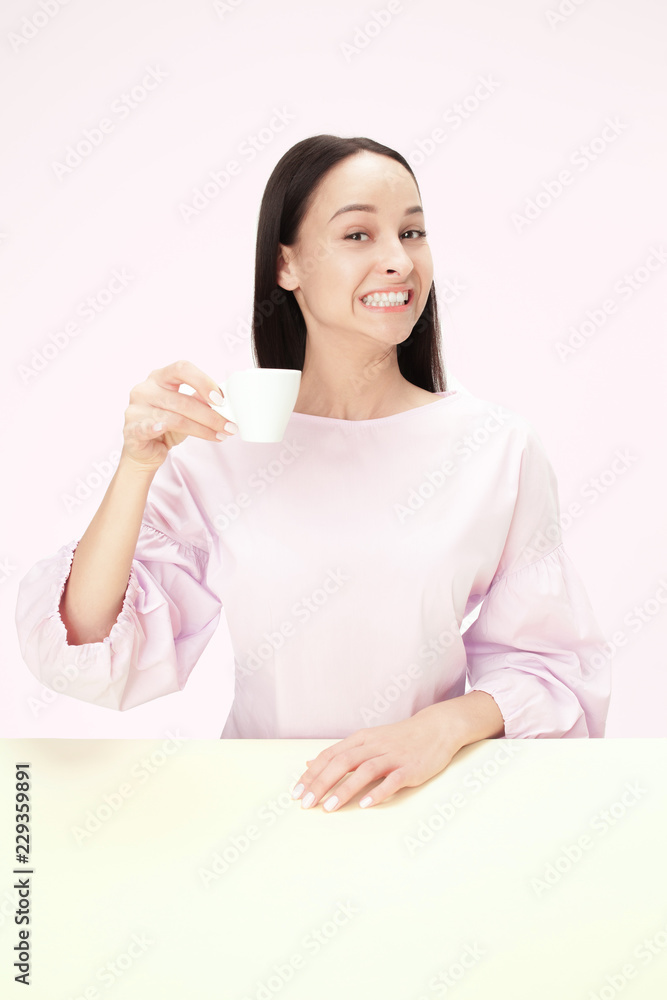Beautiful smiling woman sitting at pink studio and looking happy holding the cup of coffee in hand. Closeup toned portrait in minimalism style Beautiful smiling woman sitting at pink studio and looking happy holding the cup of coffee in hand. Closeup toned portrait in minimalism style