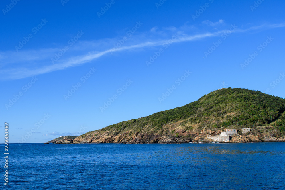Sea view of a tropical island against a cloud that follows the shape of a hill on the island. Green tropical island with the remains of an ancient fortress on the shore.