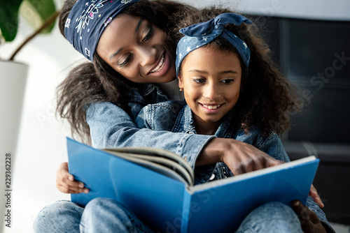 portrait of african american mother and daughter looking at family photo album at home