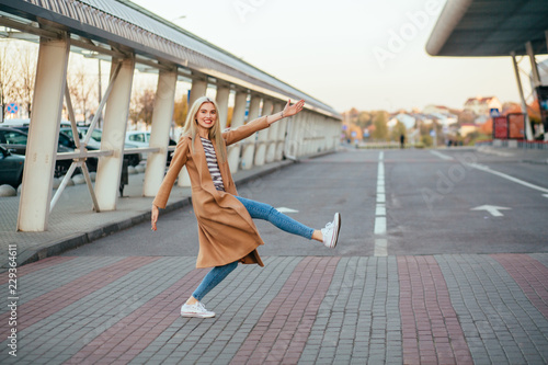 Full height of playful funny blond elegant woman in beige coat crossing the street at a pedestrian crossing.