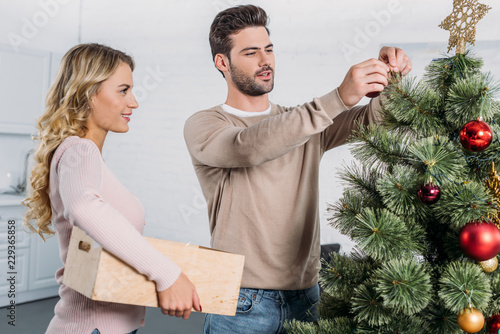 couple decorating christmas tree with baubles together at home, girlfriend holding wooden box with toys