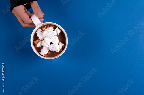 Feminine hand holding hot chocolate with marshmallow candies on blue paper background. Top view. Copy space.