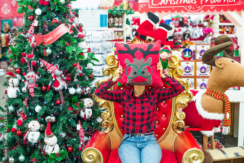 Woman in a plaid shirt holding pillow with deer instead of head and sitting on Santa Claus s throne with Christmas tree  toy deer  gift boxes. Festive market photo area in the shop  store. Copy space.
