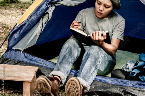 Asian young man sitting is holding a pen writing note of  letter memorize memories on book  in outside the tent. Loneliness camping in forest © Akira Kaelyn