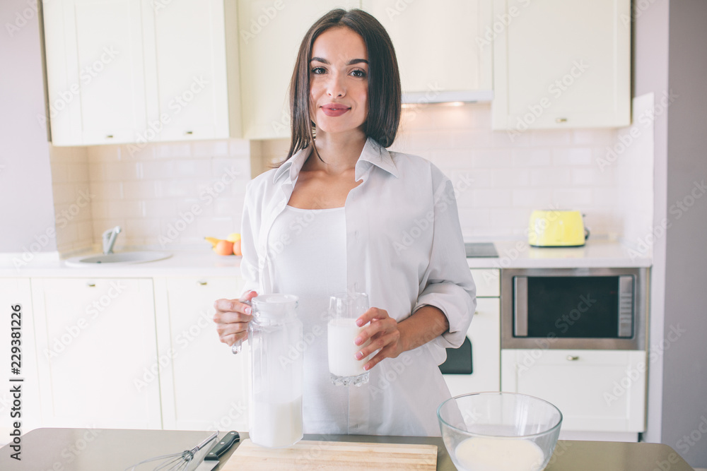 Portrait of beautiful woman stands and poses. She is looking on camera. Girl holds jar and glass of milk. She is in kitchen. Portrait of beautiful woman stands and poses. She is looking on camera. Girl holds jar and glass of milk. She is in kitchen.
