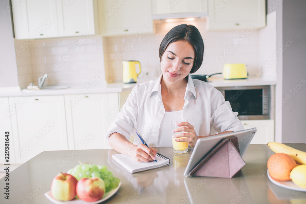 Calm and peaceful young woman sits at table and writes in notebook. She holds glass of juice in hand. There is a tablet standing on table.