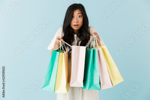Asian young shocked emotional woman isolated over blue background holding shopping bags.