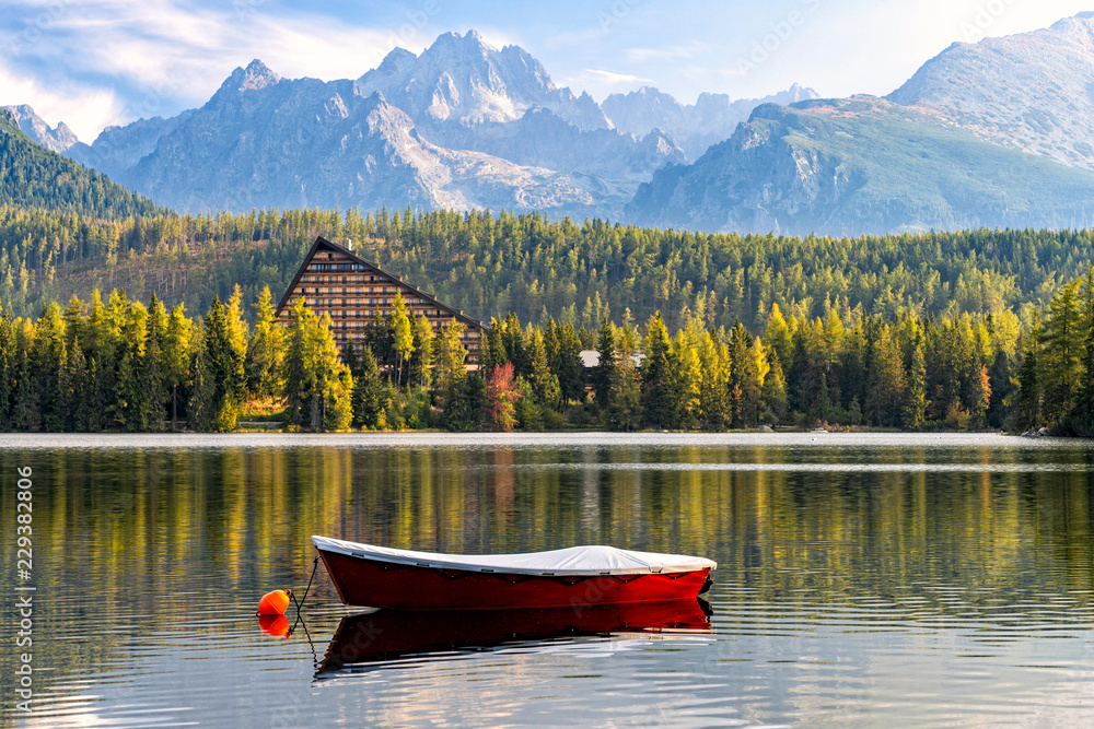 Obraz premium Peaceful mountain scene with mountain hotel next to a lake with boat. Scenic view of Strbske Pleso, High Tatras National Park, Slovakia. Focus on a boat in front.
