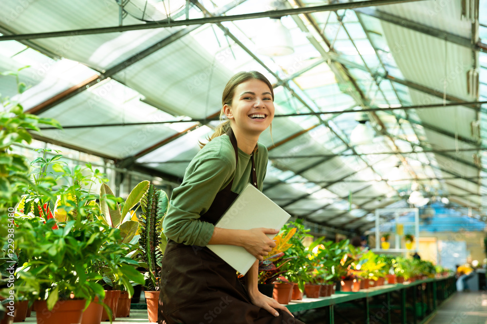 Image of joyful woman gardener smiling on camera, while standing near plants in conservatory with register in hands Image of joyful woman gardener smiling on camera, while standing near plants in conservatory with register in hands