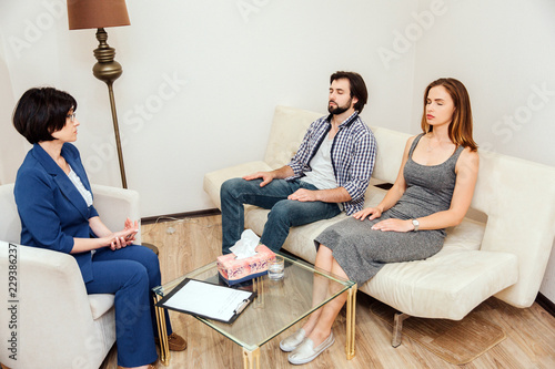 Calm and peaceful people are sitting together with their eyes closed. They are working with psychologist. Doctor is looking to young couple and talking to them.