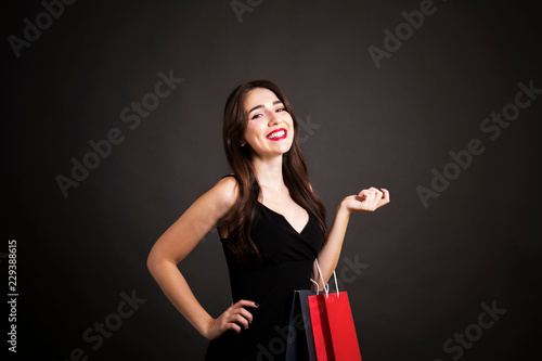 Black friday sale concept. Attractive young woman with long brunette hair, smiling, wearing sexy dress, holding red blank shopping bag over black background. Copy space, close up.
