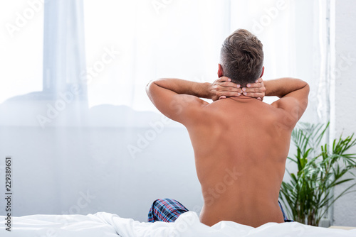 rear view of shirtless man with hands behind neck sitting on bed at home