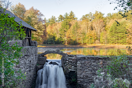 Stone wall and building with waterfall and Autumn trees reflected on Lake in upstate New York.
