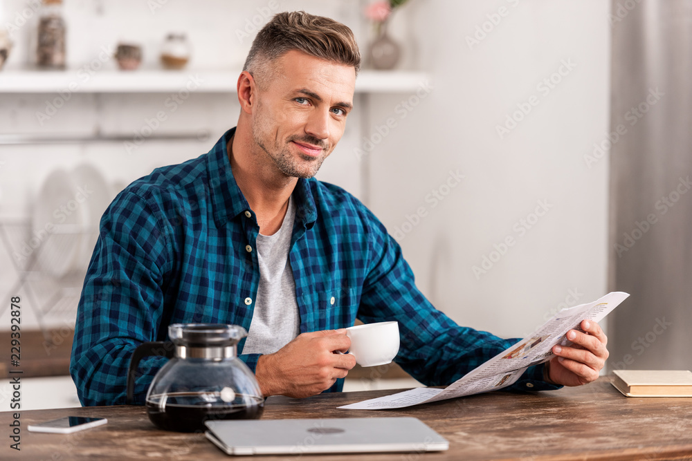 happy man with coffee cup reading newspaper at table with laptop in kitchen at home happy man with coffee cup reading newspaper at table with laptop in kitchen at home