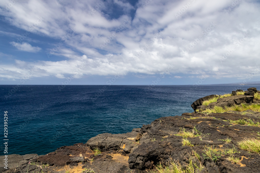 View from South Point on the Big Island of Hawaii. This is the southernmost point in the United States. Past the lava rock cliff is the deep blue pacific Ocean; blue sky and clouds in the distance. 