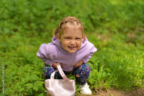 A little girl in a purple jacket sitting on the grass makes faces.