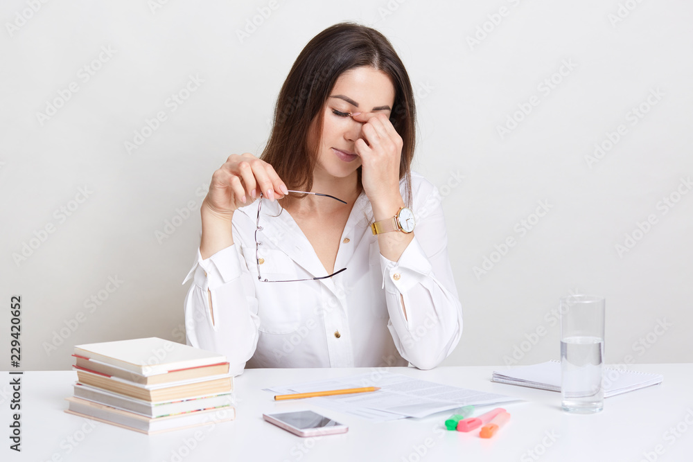 Tired young lady tries to concentrate on work, keeps hands on nose, holds spectacles, dressed in white formal shirt, poses at work space with documents and books, isolated over white background Tired young lady tries to concentrate on work, keeps hands on nose, holds spectacles, dressed in white formal shirt, poses at work space with documents and books, isolated over white background