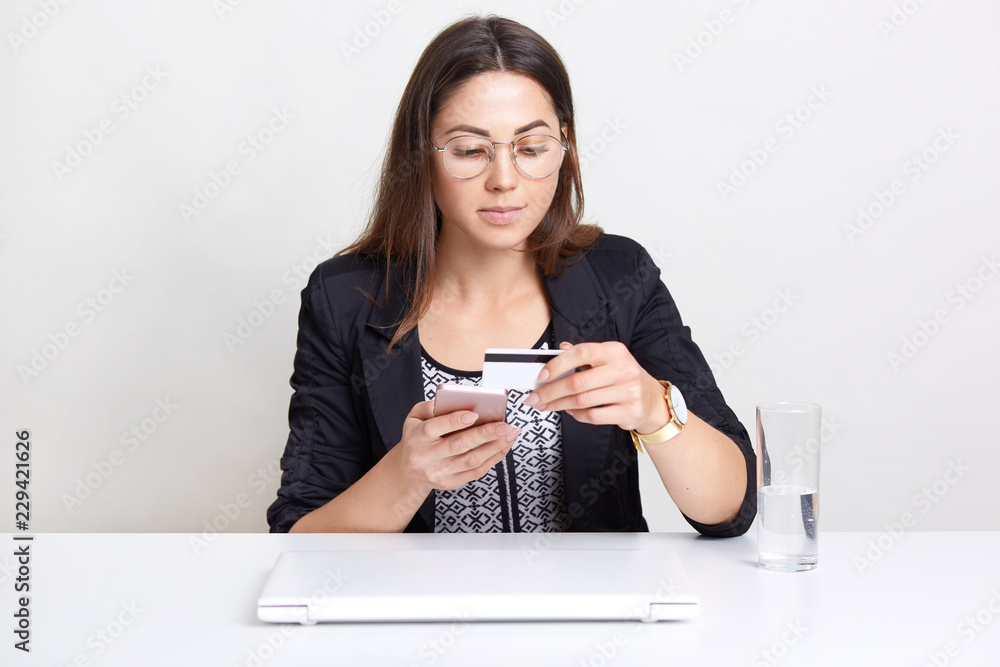 Pretty woman holds plastic credit card, does verification of banking data, has online shopping, uses modern technologies, dressed in formal outfit, isolated over white background. Technology