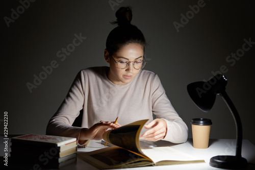 Horizontal shot of focused female scientist turns over pages in book, searches necessary information, drinks hot coffee, wears glasses, uses reading lamp, sits at work space alone. Reading concept photo