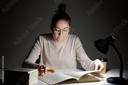 Photo of focused woman has hair knot, has serious clever expression in textbook, sits at desk with lamp, busy with exam preparation, poses indoor. Dark colours. People, education and learning. photo