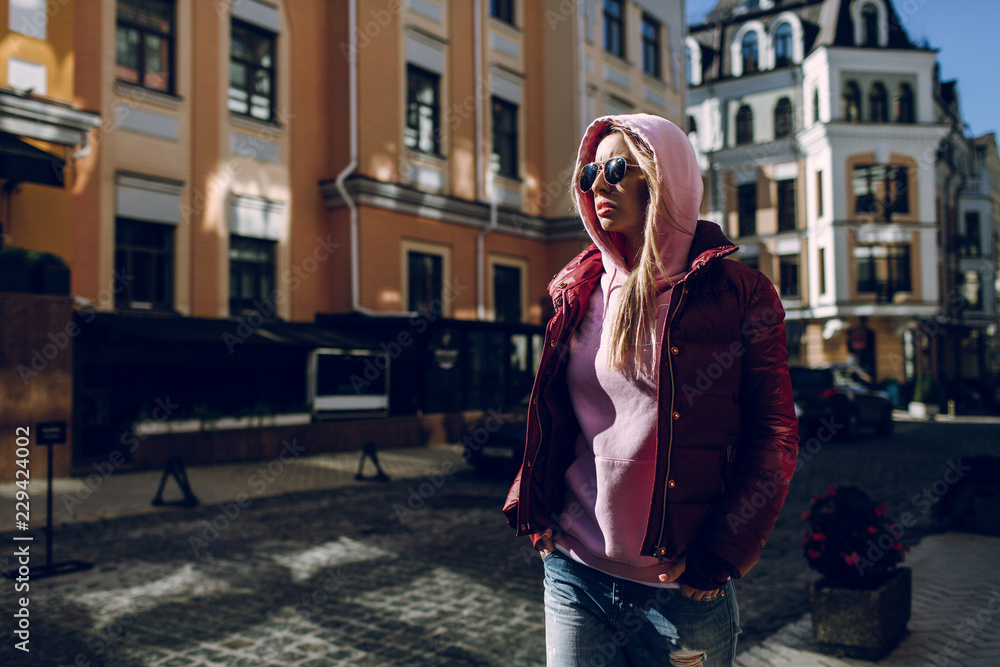 Fototapeta premium Street fashion concept - portrait of a beautiful girl sitting outside, blue jeans, sunglasses, hard lights, red jacket, autumn weather