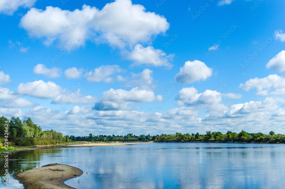 The view from the shore to the wide spilled river with shallow areas under the blue sky with beautiful white clouds. Belarusian Polesie. Pripyat National Park