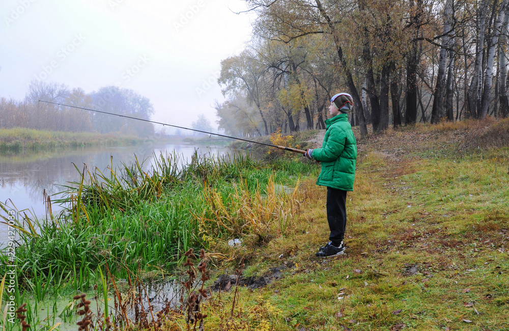 A child of 9 years old in a green jacket and with a fishing rod in his hands during the morning autumn fishing, river fog.